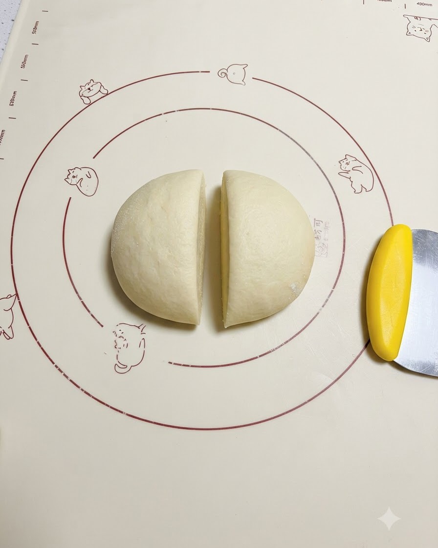 Proofed dough cut evenly in half on a patterned silicone mat next to a yellow dough scraper.
