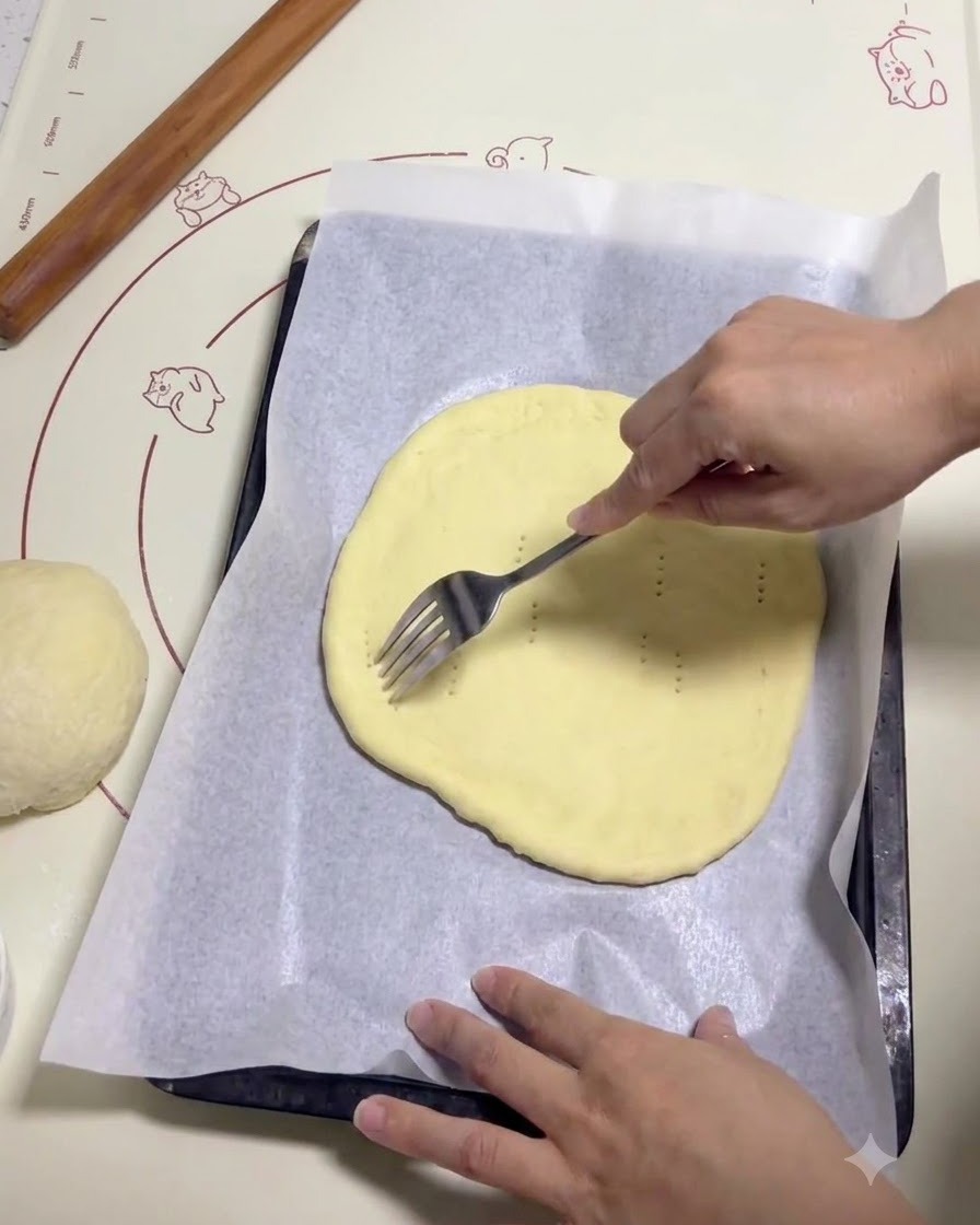 A hand using a metal fork to poke small holes across a flat, round piece of pizza dough on parchment paper.