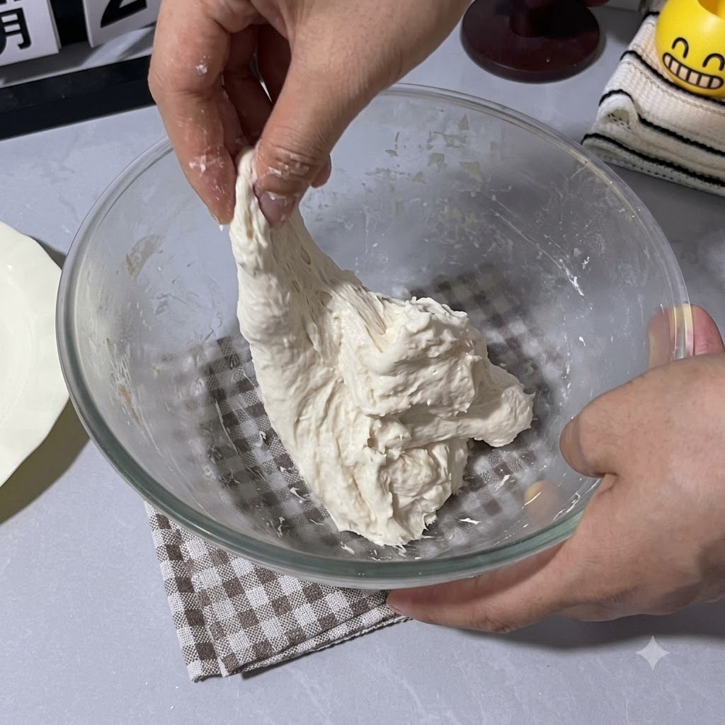 A hand pulling and folding the sticky dough upward inside the glass bowl.