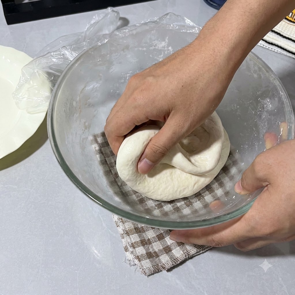 Hands spraying with water folding the dough inside the glass bowl covered with plastic wrap.