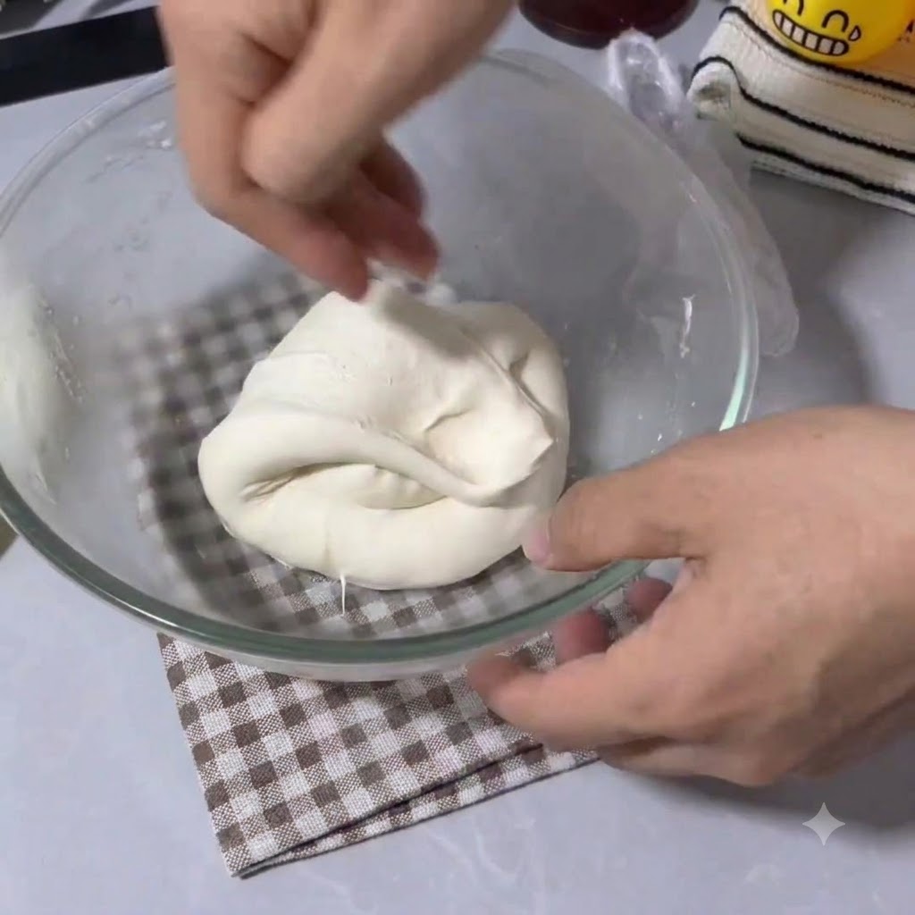 Hands folding and stretching elastic pizza dough inside a clear glass mixing bowl.