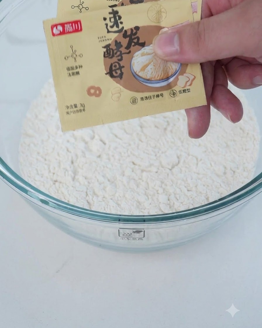 Dry and wet ingredients including flour and yeast being added into a clear mixing bowl.