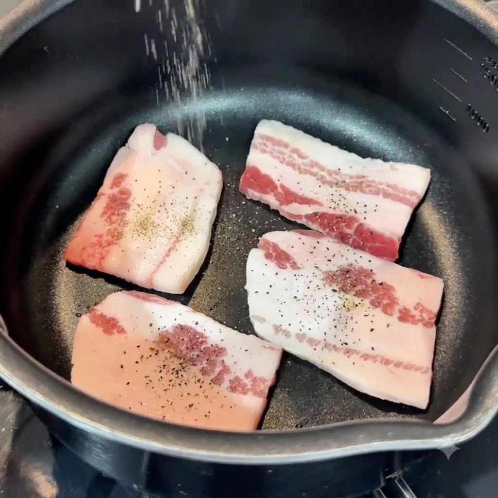 Salt and pepper being sprinkled from above onto pork belly slices inside a pan.