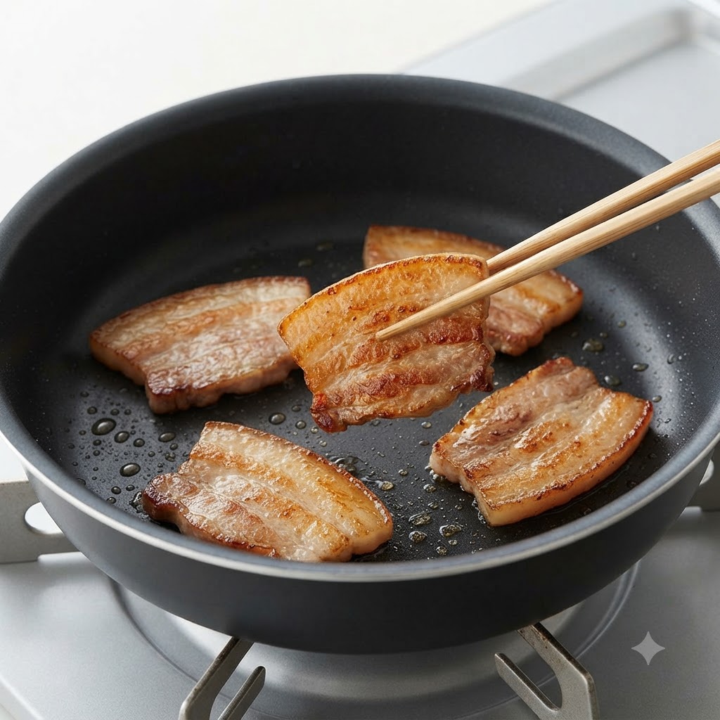 A pair of chopsticks flipping a browned and seared slice of pork belly in a pan.