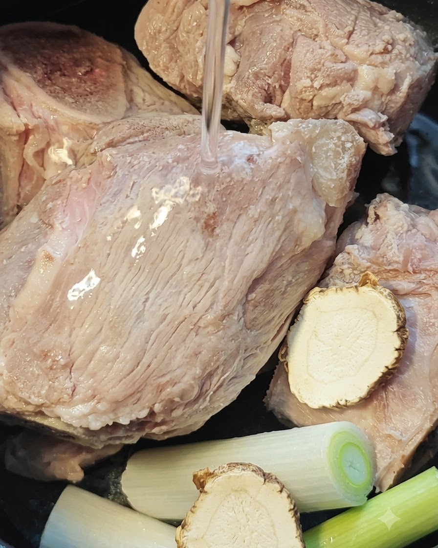 Water being poured over pork bones, angelica root, and scallions inside a rice cooker pot.