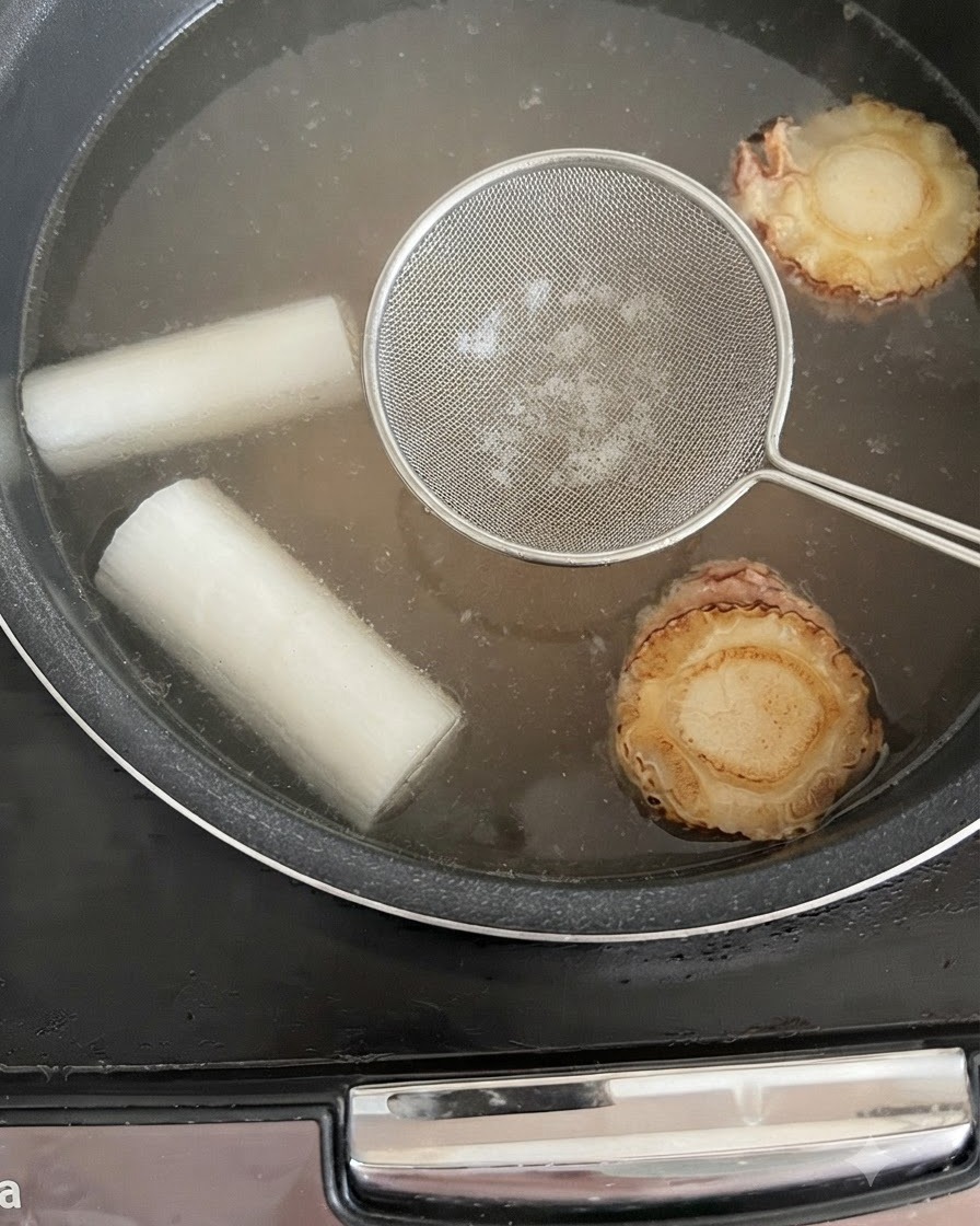 A skimmer tool held over the simmering broth in the rice cooker to remove impurities.
