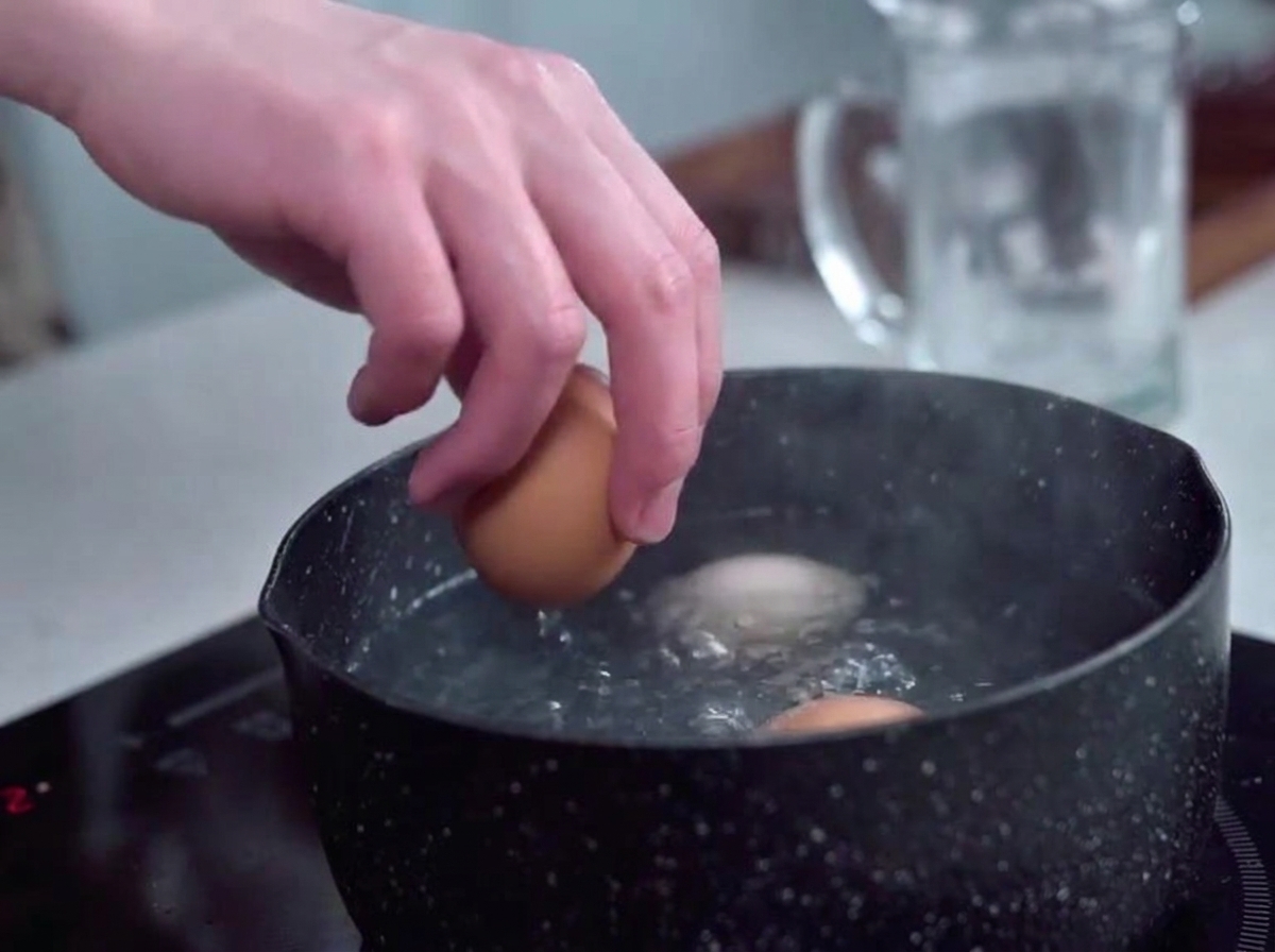 Eggs being gently placed into a pot of bubbling boiling water using a hand.