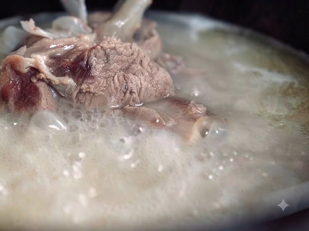 A thick, milky, and opaque pork bone broth boiling vigorously in a pot, with pieces of meat and bone visible.