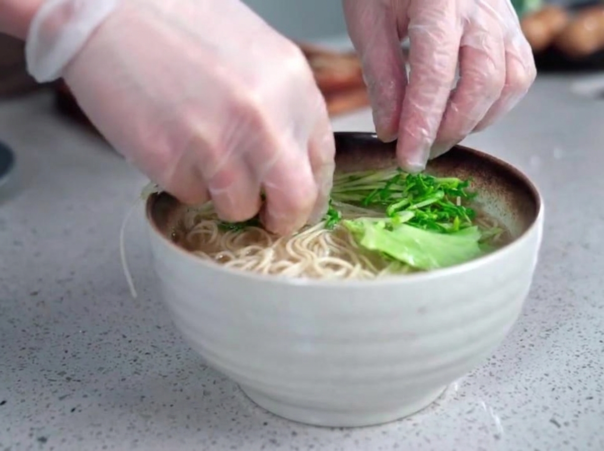 Gloved hands carefully placing fresh green leafy vegetables into a steaming bowl of ramen noodles and opaque broth.
