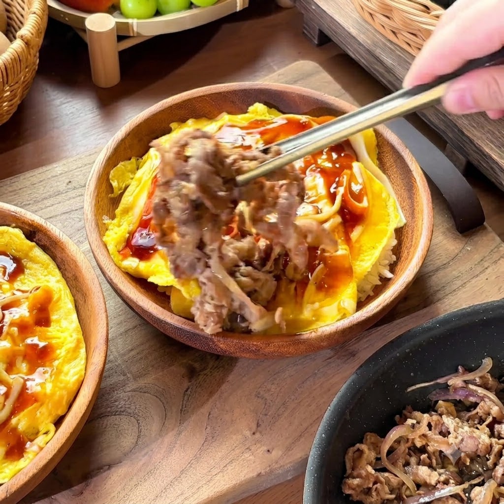 Chopsticks lifting a portion of cooked beef and onions to place onto a bowl of rice and eggs covered in sauce.