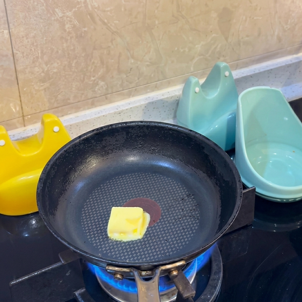 A yellow cube of butter melting in the center of a black non-stick frying pan over a gas flame.