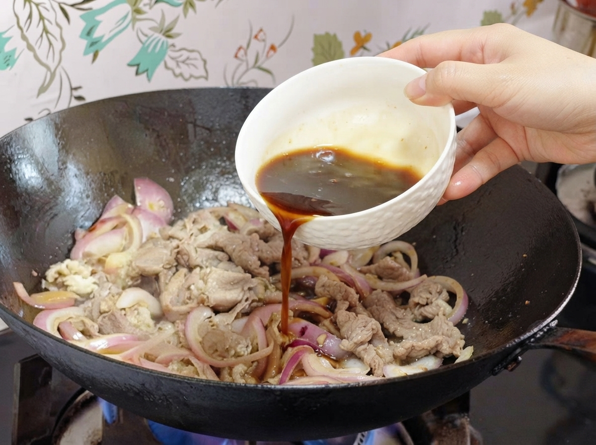 Sauce being poured from a small white bowl into a wok with onions and beef slices.