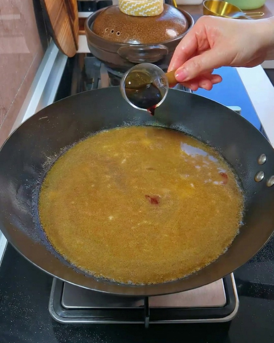 A hand pouring dark soy sauce from a small measuring glass into a wok filled with broth.