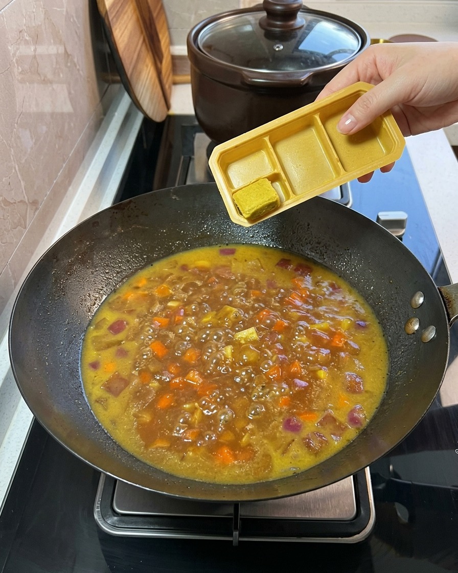 Solid curry cubes being added from a plastic tray into the simmering vegetable and broth mixture.