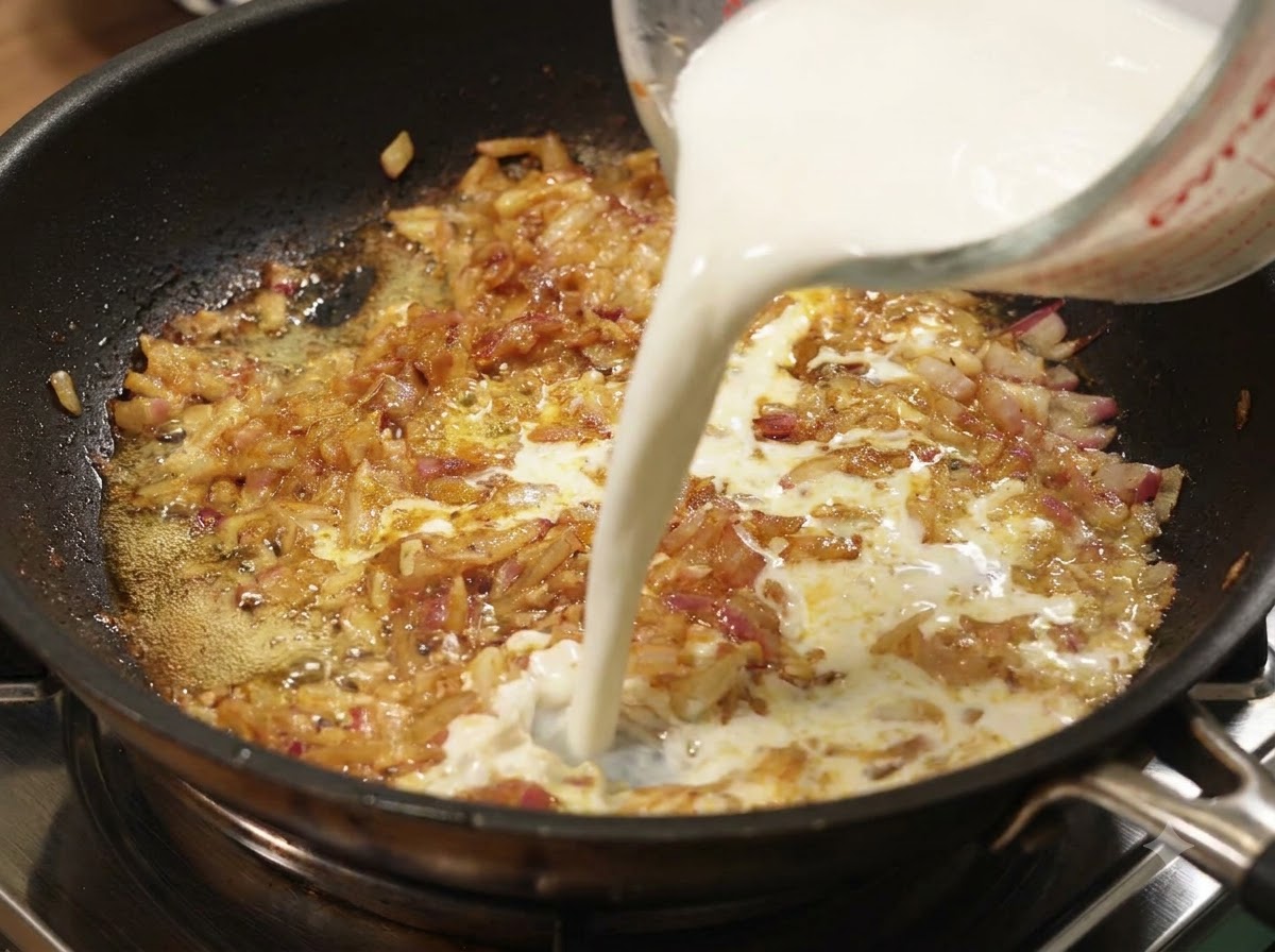 Milk being poured from a measuring cup into a pan of simmering onions and curry.