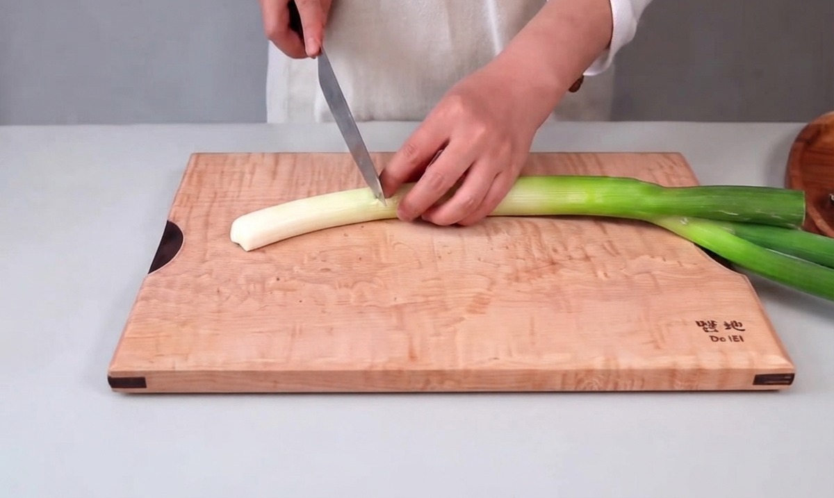 Hands holding a kitchen knife slicing a large green onion on a wooden cutting board.