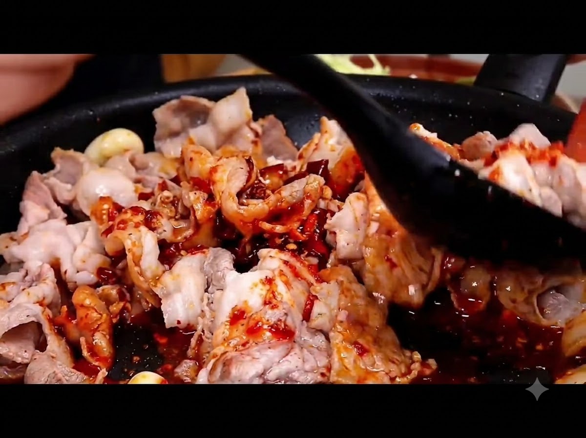 A black spatula tossing and mixing spicy red sauce into pork belly slices in a hot skillet.