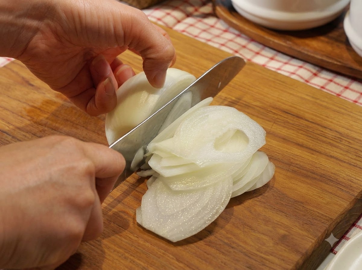 An onion being carefully sliced into thin half-moons on a wooden cutting board with a chefs knife.