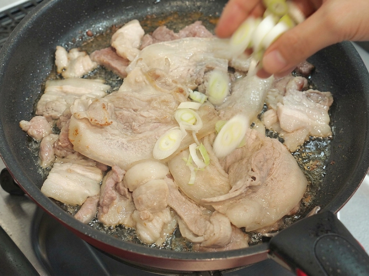 Chopped green onions being dropped into a pan containing cooking pork belly slices.
