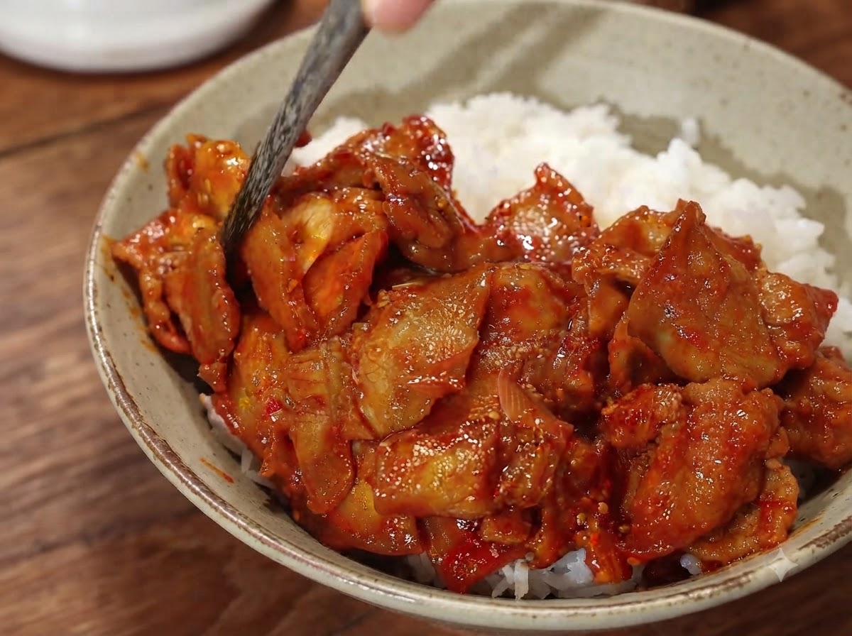 A close-up of a spicy pork rice bowl being finished with soaked sliced onions and chopsticks adjusting the toppings.