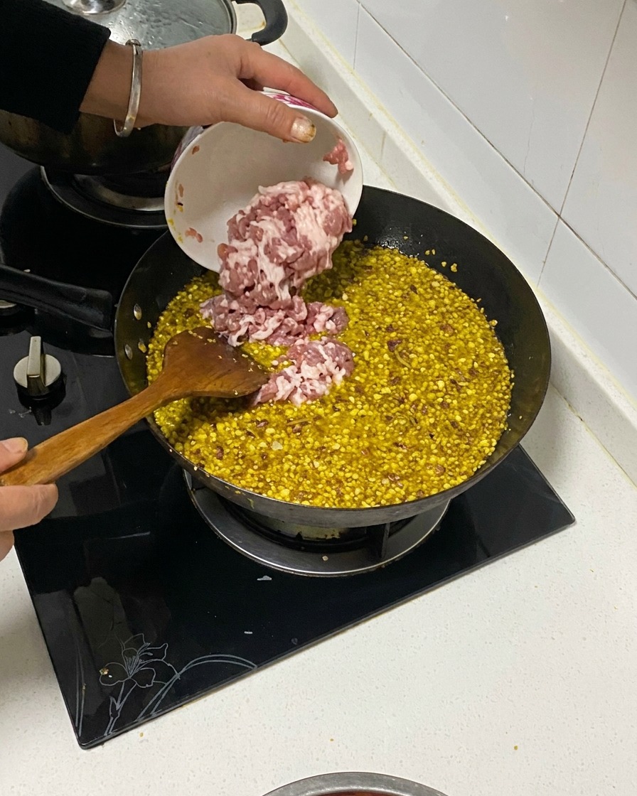 Diced pork belly being poured from a small bowl into a wok containing fried aromatics and oil.