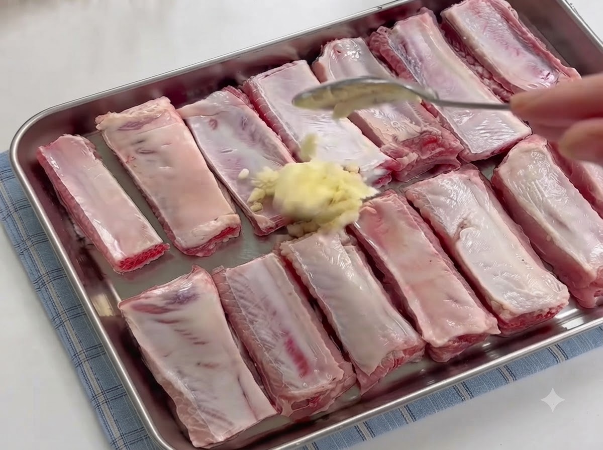 A spoon adding a mound of freshly minced garlic onto raw spare ribs in a tray.
