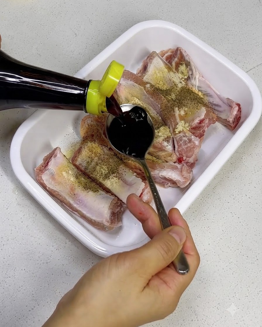 Pouring dark soy sauce from a bottle onto raw pork ribs in a white tray.