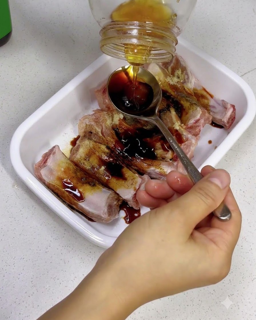 Hand holding a spoon to catch golden honey being poured from a glass jar over seasoned ribs.