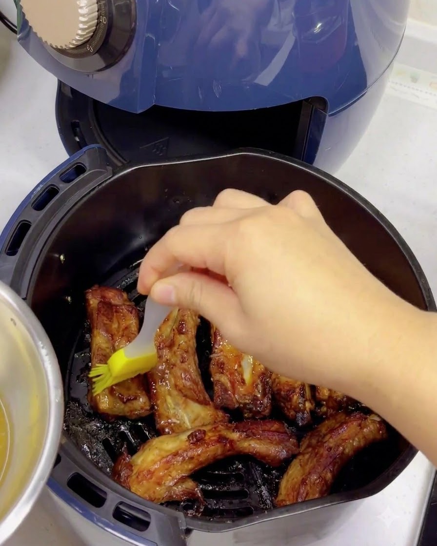 A yellow silicone brush applying a honey water glaze over cooked, browned pork ribs inside an air fryer basket.
