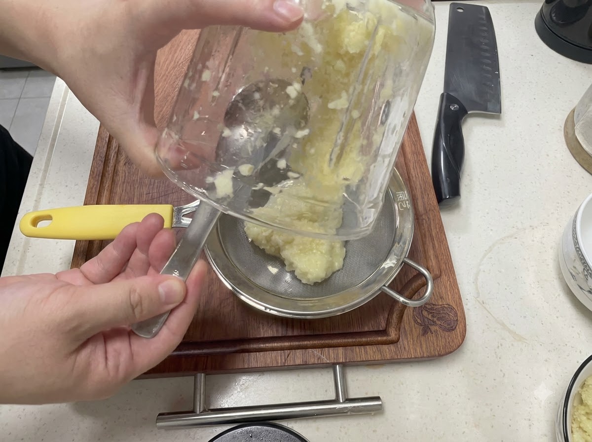 Blended garlic being poured from a glass container into a fine-mesh strainer over a metal bowl.