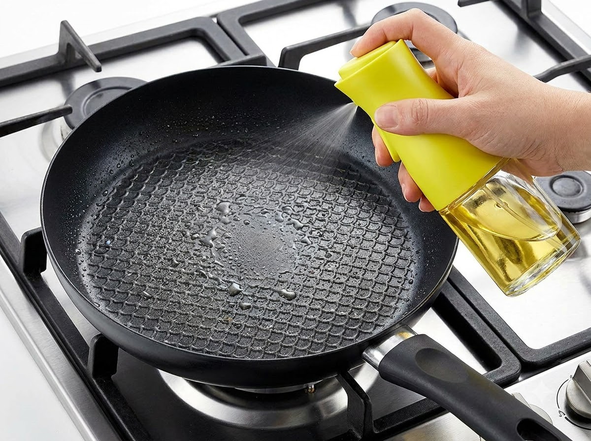 A hand spraying cooking oil into a black frying pan set over a stovetop burner.