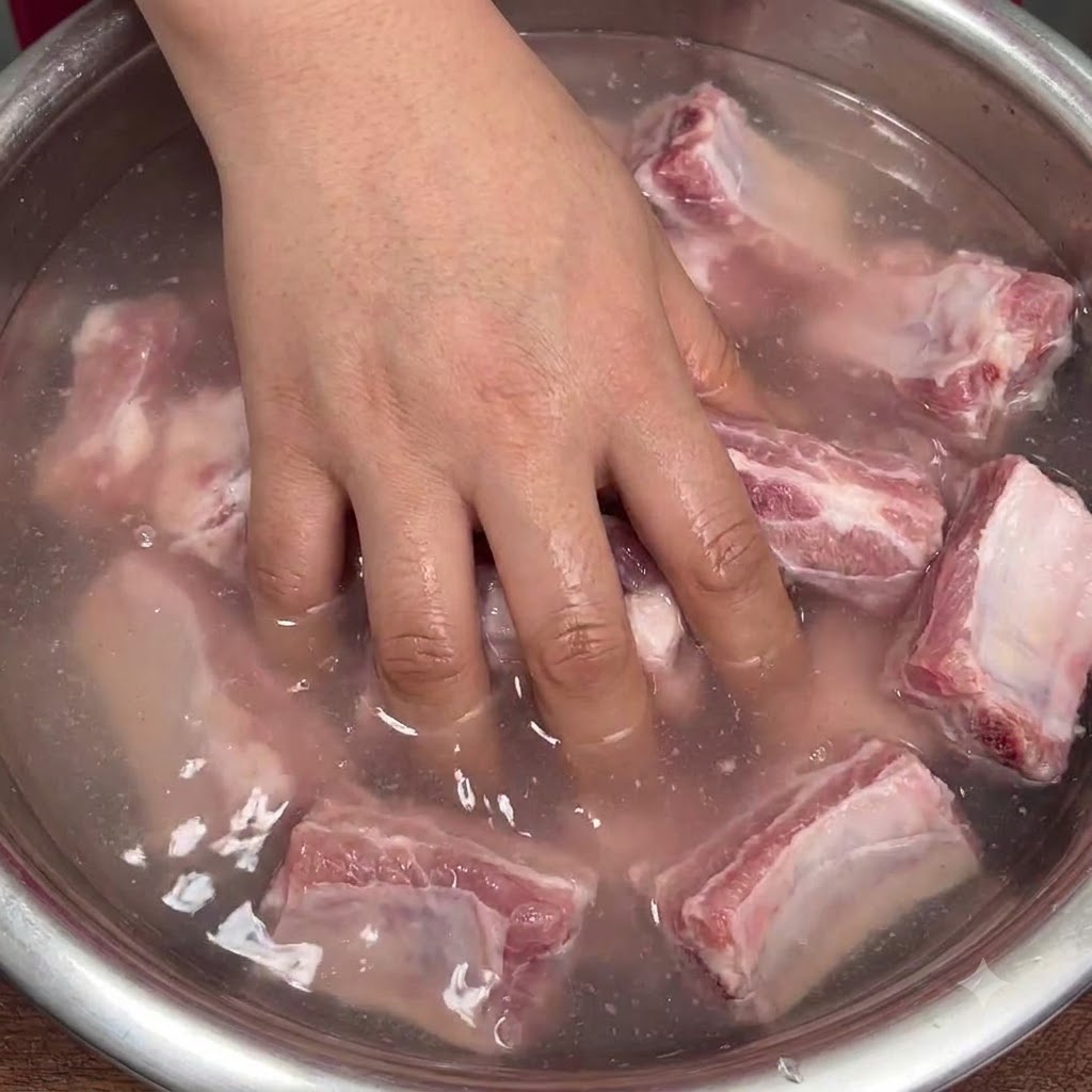 A hand vigorously mixing raw pork ribs in a bowl of cloudy water to distribute the baking soda.