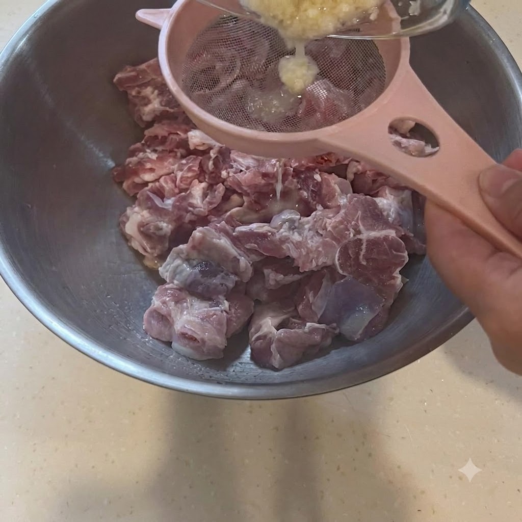 Garlic paste being strained through a pink sieve directly over a large bowl of raw pork pieces.