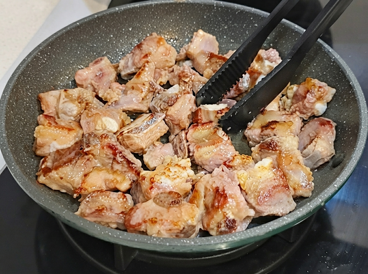 Pork ribs being flipped in a non-stick frying pan using black tongs.