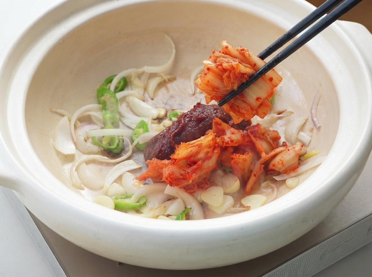 Korean chili paste and kimchi being added to the pot of aromatics using chopsticks.