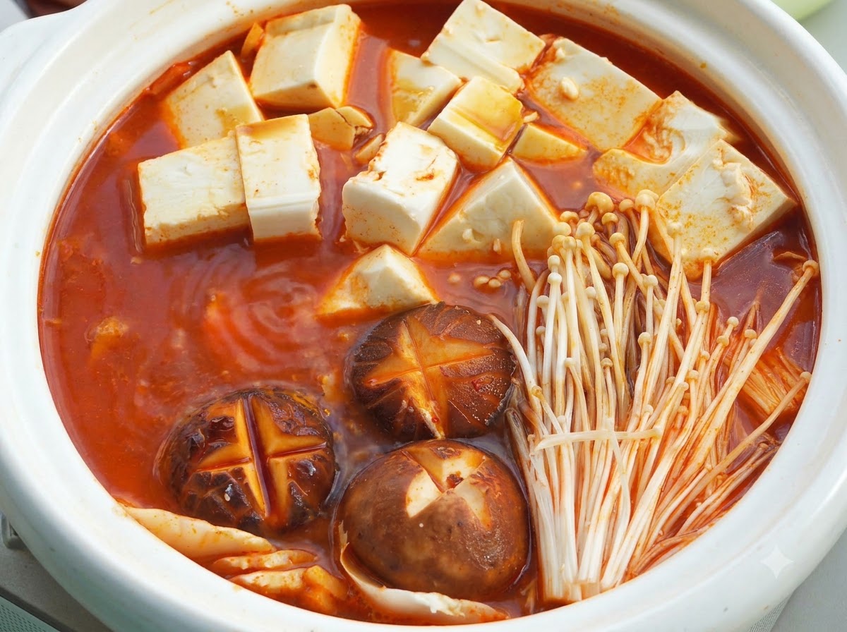 A white ceramic hot pot filled with simmering red soup base, soft tofu cubes, shiitake mushrooms with cross cuts, and enoki mushrooms under a lid.
