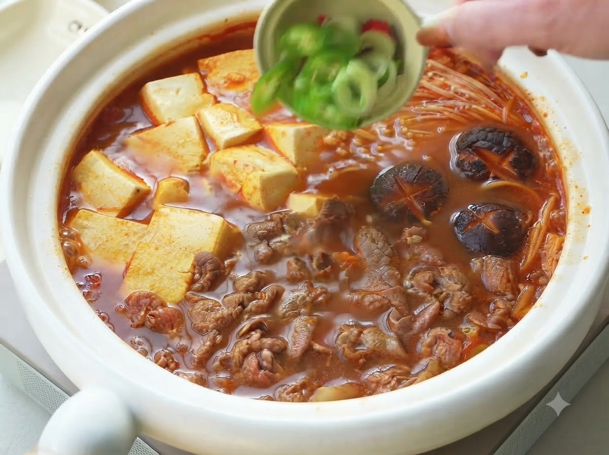 Green onions and sliced chili peppers being poured from a small bowl as a garnish over the cooked Korean beef and tofu stew.