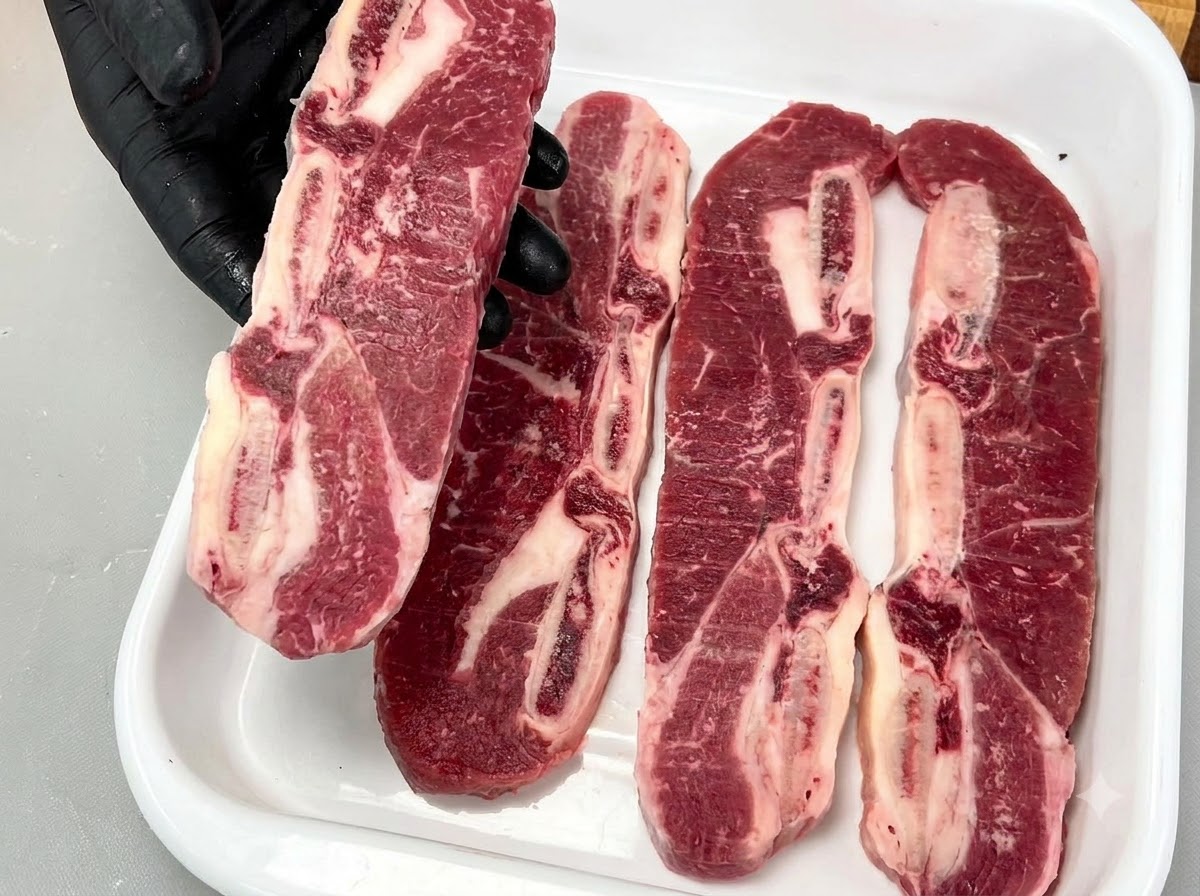 A gloved hand holding up a raw bone-in LA short rib, displaying its rich marbling and bone structure.