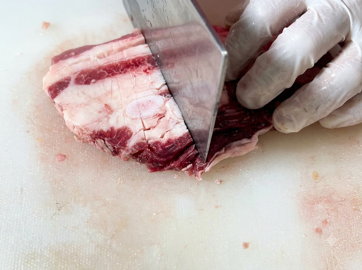 Raw beef short ribs being sliced into uniform rectangular pieces on a cutting board using a large knife.