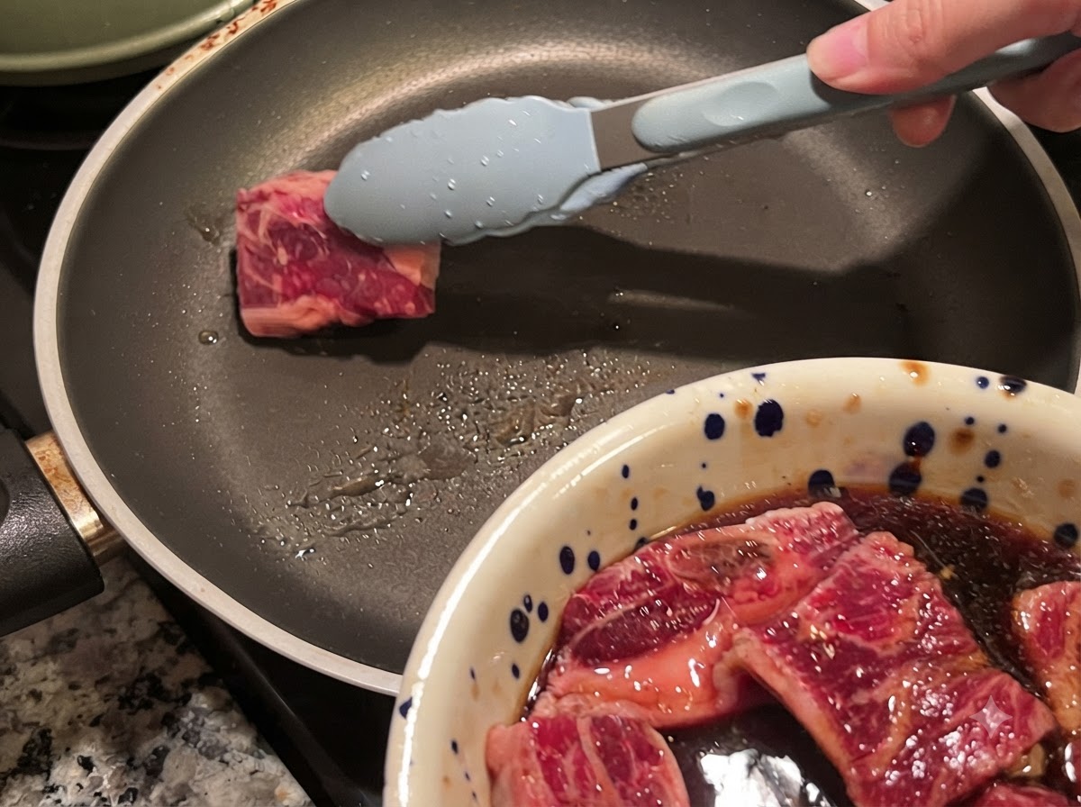 Tongs placing a piece of marinated beef short rib into a hot skillet.