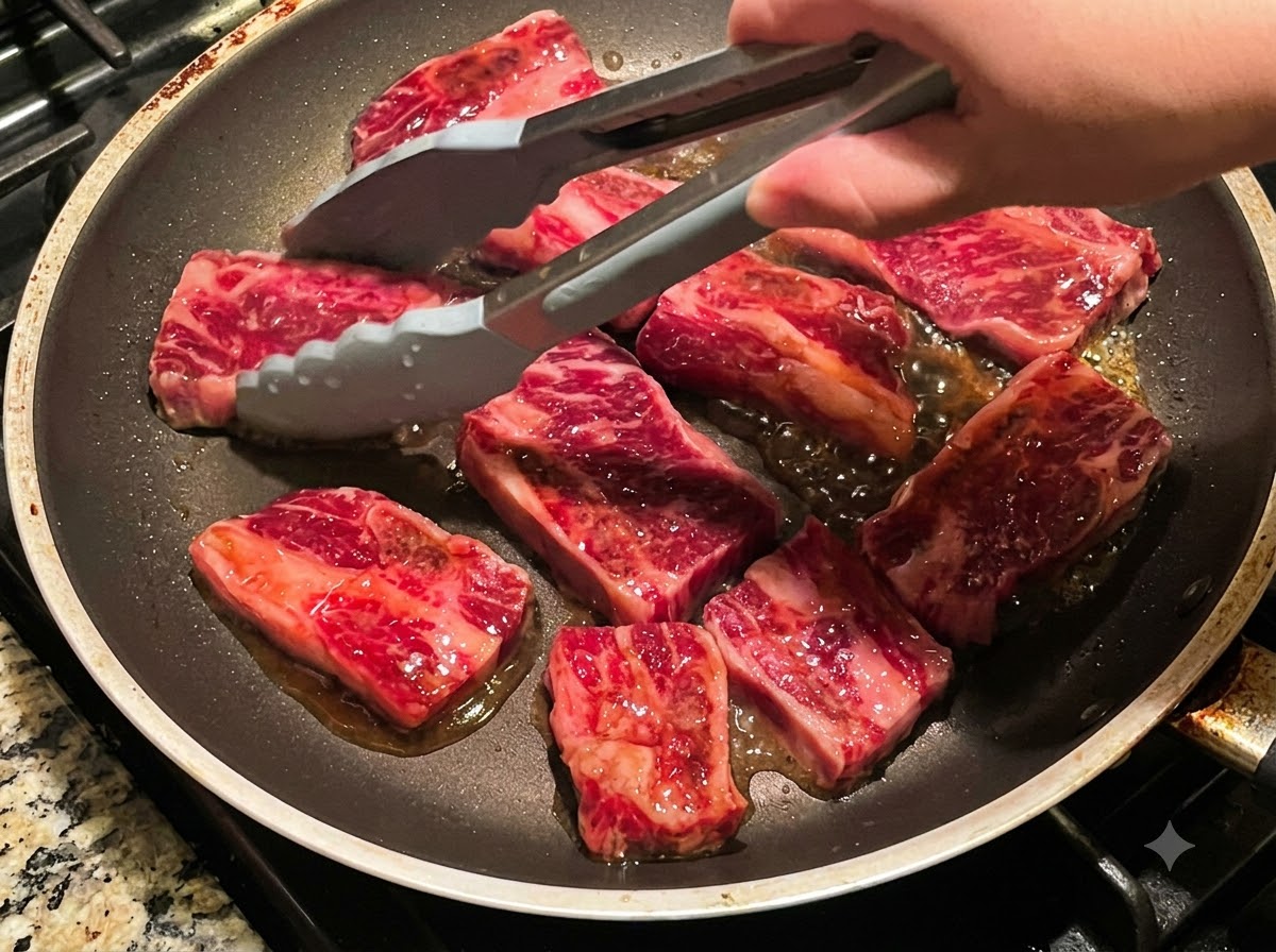 Tongs flipping multiple pieces of beef short ribs frying in a pan.