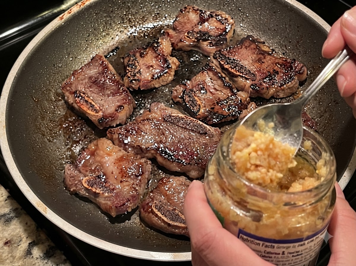 Minced garlic being spooned from a jar into a hot skillet with browning beef short ribs and onionscite: 1.