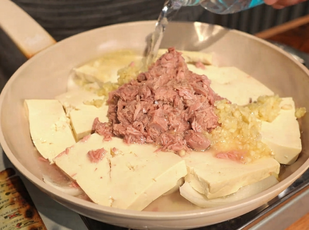 Clear water being poured from a bottle into a pan containing layered tofu, onions, garlic, and canned tuna.