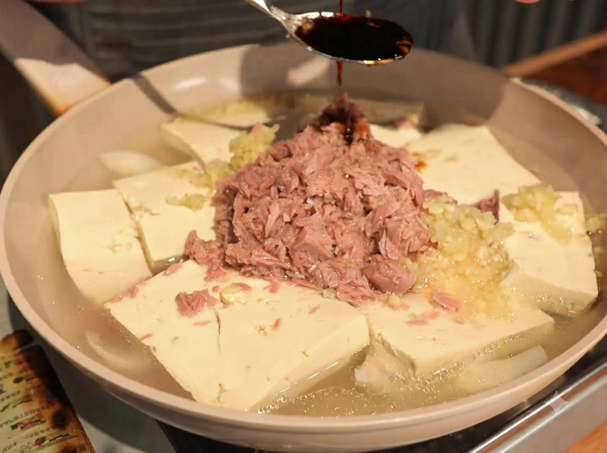 Dark soy sauce being drizzled from a spoon onto the center of canned tuna and tofu pieces.
