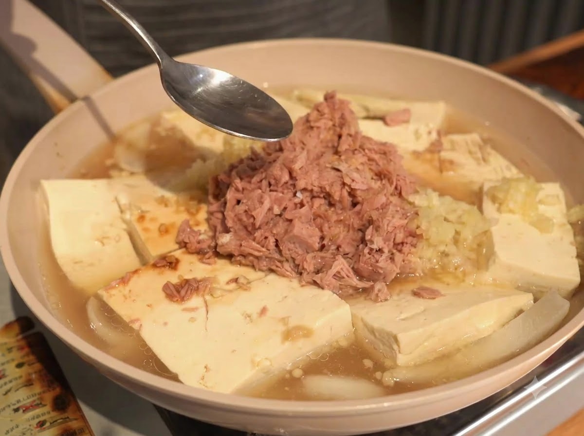 A spoon held over the pan containing tuna, tofu, and liquid broth during the seasoning process.
