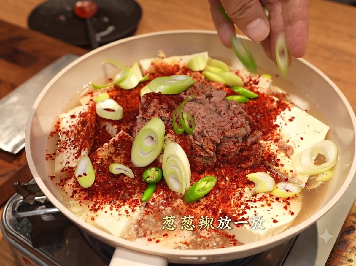 Hands scattering sliced green onions and green chili peppers over a pan covered in coarse red chili flakes, tofu, and tuna.