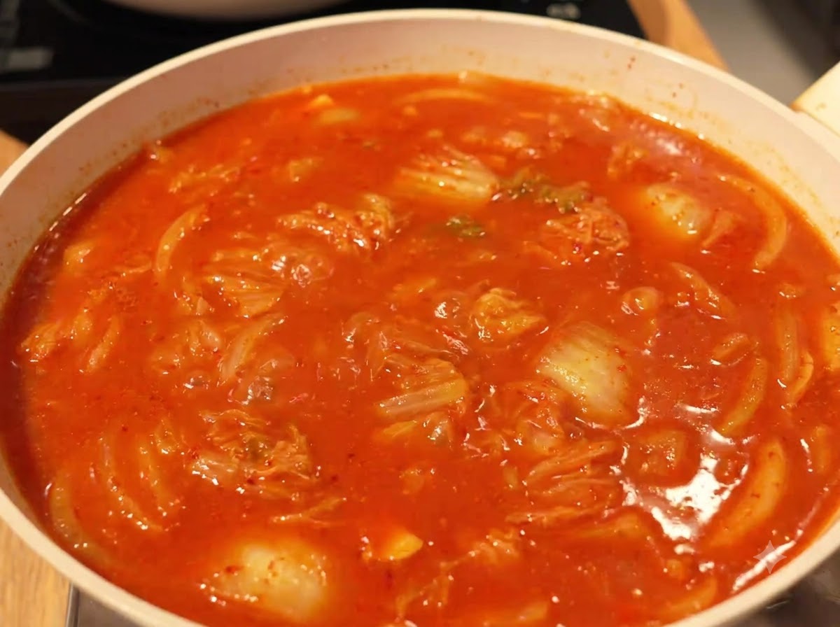 A rich, red, bubbling broth simmering with kimchi and ingredients in a pan on the stove.