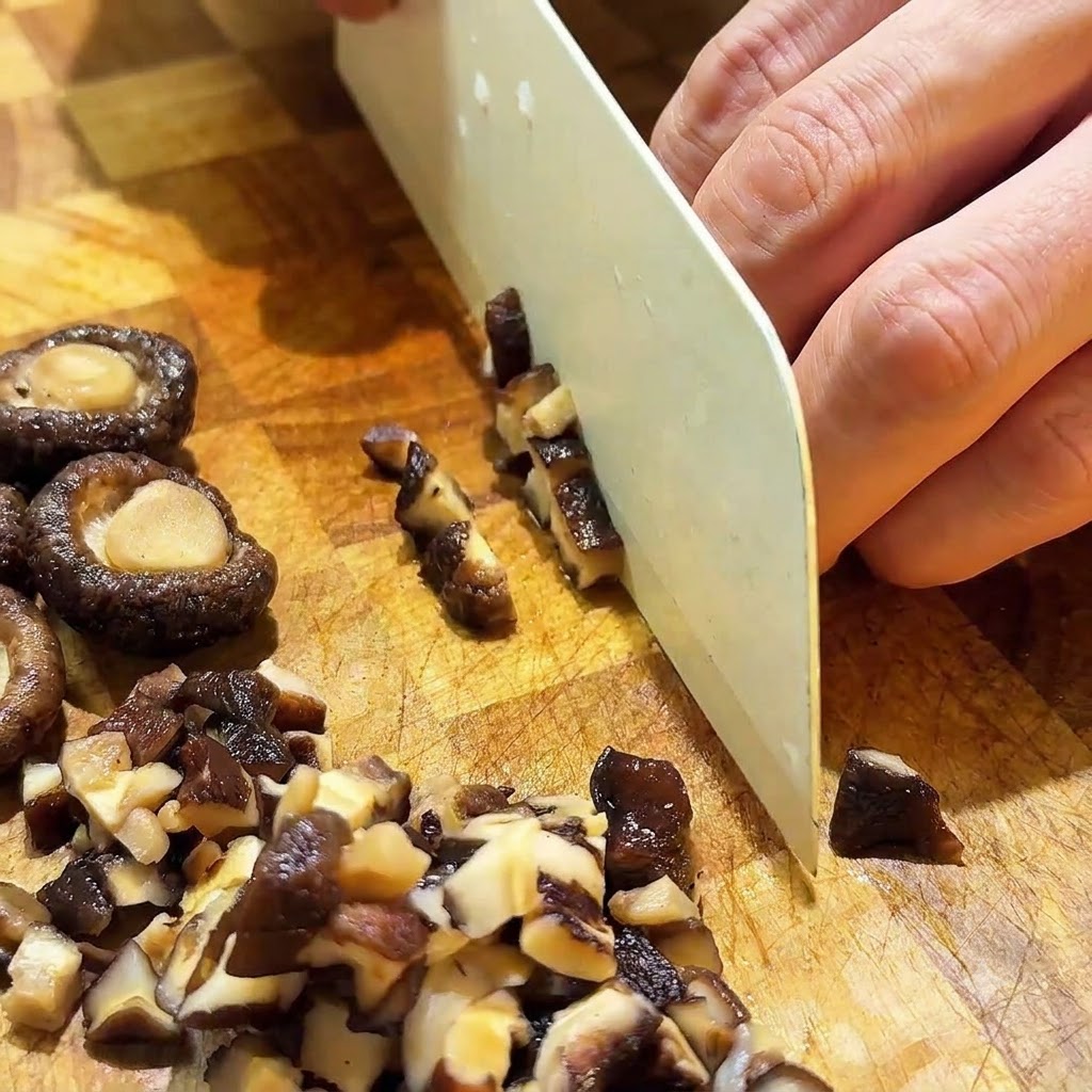 Rehydrated shiitake mushrooms being sliced into small cubes with a white knife on a wooden board.