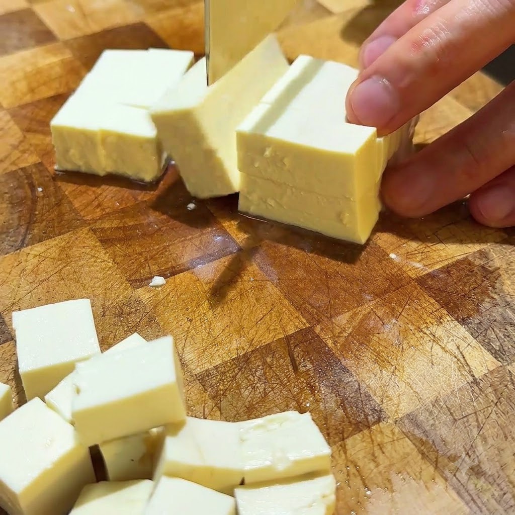 Silken tofu being cut into neat small cubes on a wooden cutting surface.