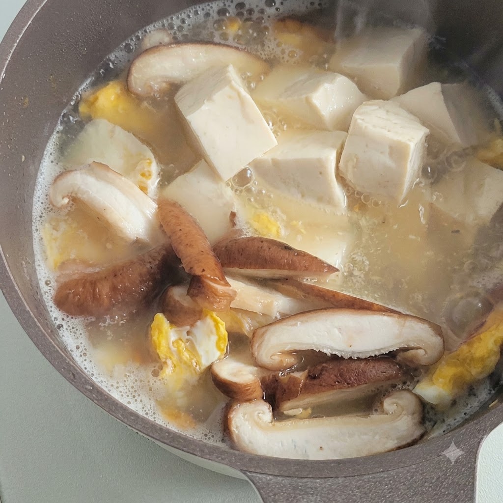 Cubes of soft white tofu simmering in a savory broth alongside sliced mushrooms and fried egg pieces.